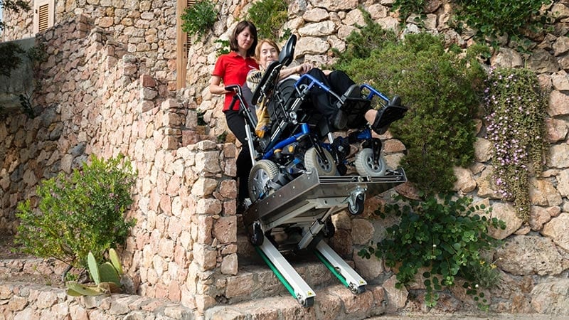 An elderly disabled person in a wheelchair is transported up an outdoor stone with the electric crawler wheelchair stair climber Domino People